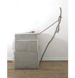 A sculpture composed of three rectangular, textured forms, a stick tied with a rope that is leaning away from them on the right side, and a piece of light fabric draping off the left side.