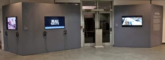 Grey walls with 3 screens showing videos, a set of doors in the center, and illegible gallery wall text on the right.