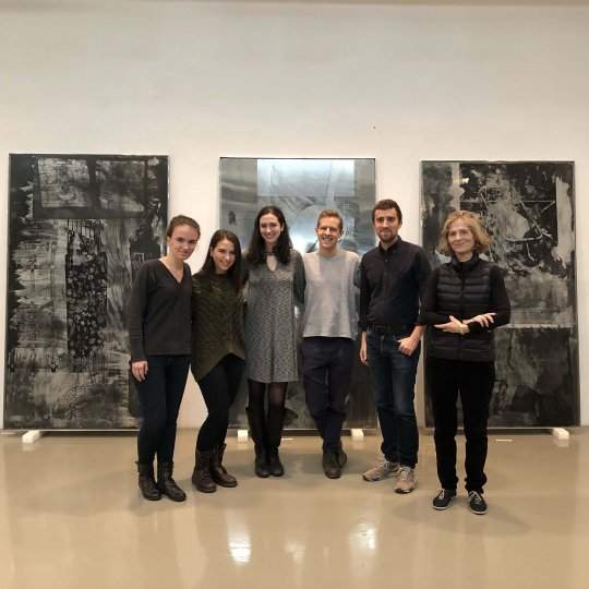 A group of six people wearing black, grey, and dark blue clothing, standing in front of three large artworks. The artworks contain black and grey silkscreened imagery on mirrored and brushed aluminum.