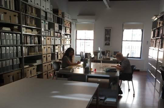 Two people on opposite sides of a large table studying documents. There is a wall of shelves full of boxes on the left, another table in the foreground, and two windows in the background.