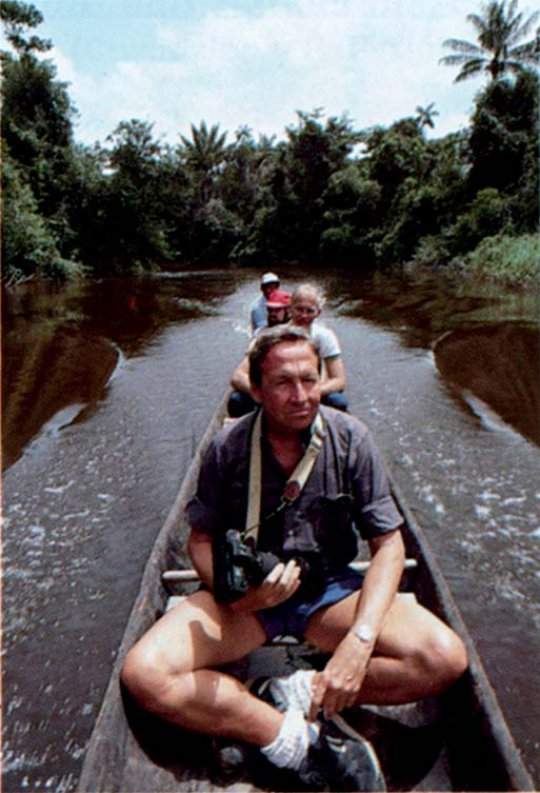 Robert Rauschenberg sits in a canoe, with a camera in his hands. Three people sit behind him. The canoe is in the middle of a river, surrounded by lush rainforest.