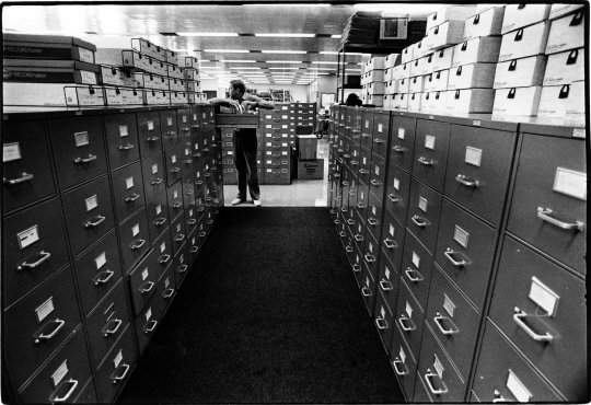Robert Rauschenberg in a room full of file cabinets and boxes, leaning on an open file cabinet drawer and looking off to the side.