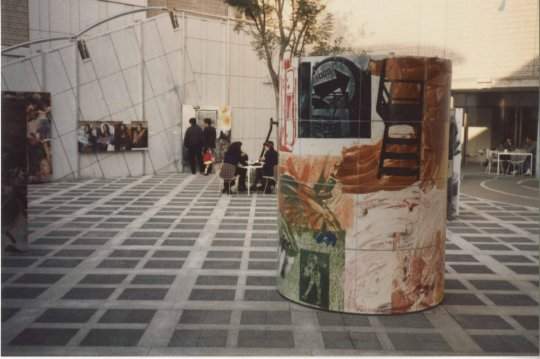 A large, colorful, cylindrical sculpture sits at the center of an outdoor courtyard. In the background, museum visitors sit at tables, while others look at artworks that hang on the courtyard walls.