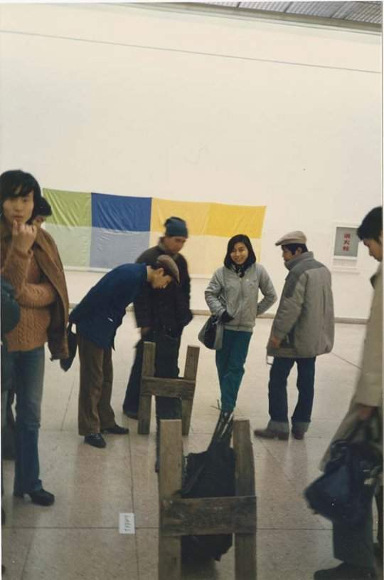 A group of people look down at a sculpture made of wood and tire tread that sits on the floor. A green, blue, and yellow fabric artwork is visible hanging on the wall in the background.