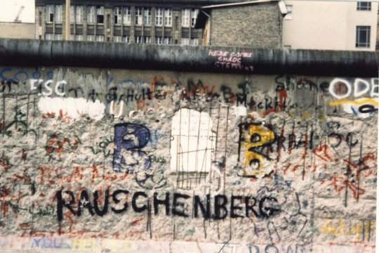 A view of a small section of the Berlin Wall. Among the largely illegible, multi-color graffiti is the name "Rauschenberg" in black, uppercase letters.