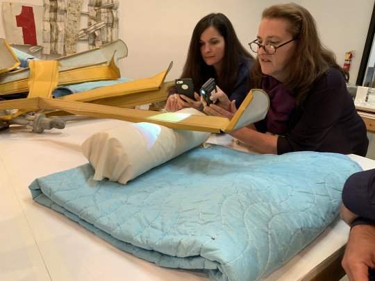 Two people examining part of a yellow, metal artwork. One is holding a shining a light, and the other is holding up a smartphone. The artwork is propped up on a white cushion and blue blanket.